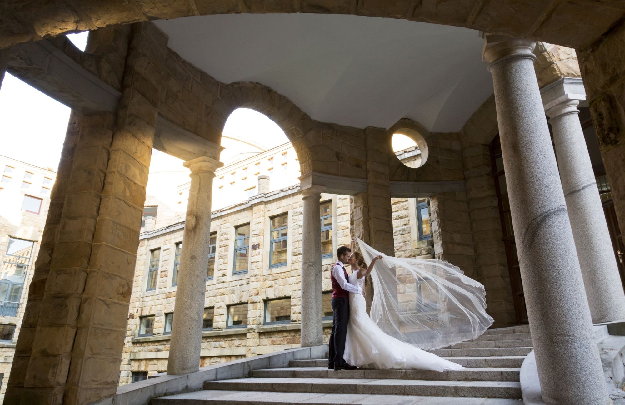 Pareja de novios bajo un pórtico de columnas de piedra, besándose con el velo al viento