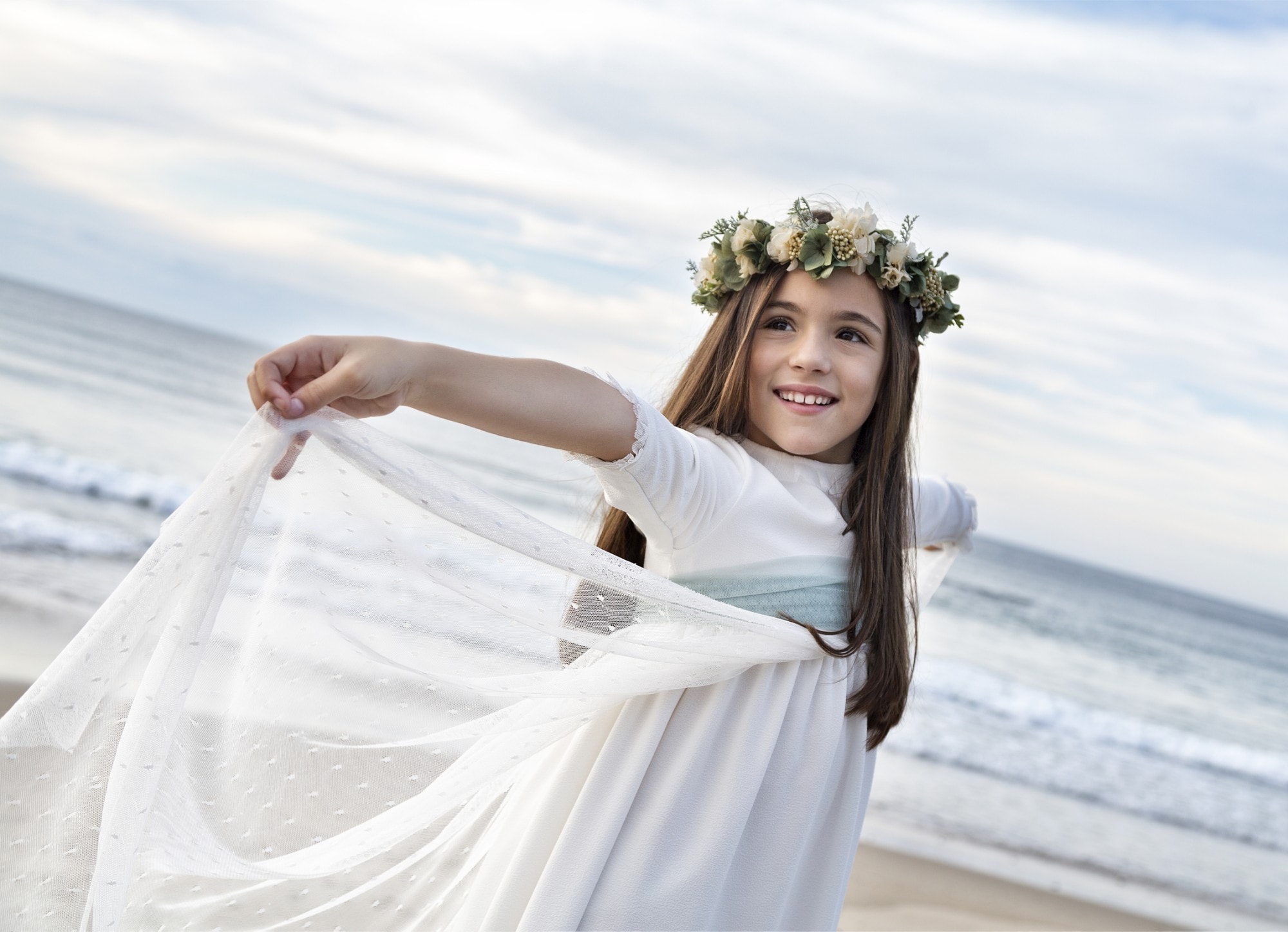 Niña de comunión en la playa al atardecer, con corona de flores y un velo blanco